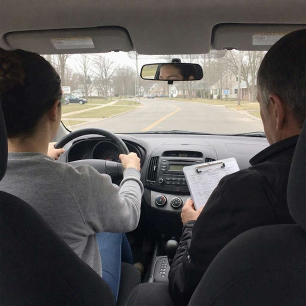 A learner driver at the wheel during a road test while the examiner marks a clipboard in the passenger seat